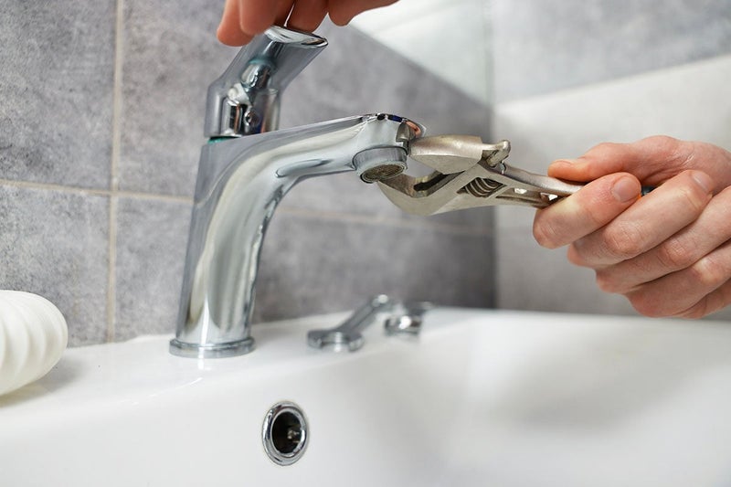 Hands using an adjustable wrench to repair a chrome faucet on a white sink.