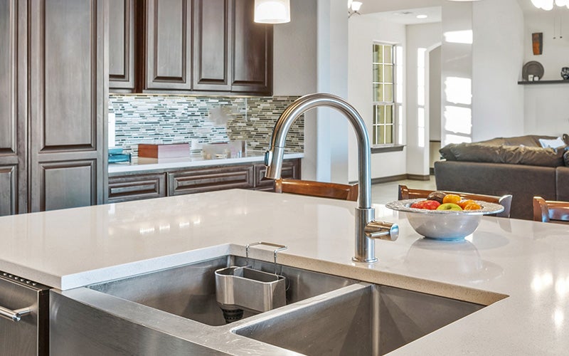A modern kitchen island with a double sink, chrome faucet, and bowl of fruit, with dark cabinets and living room beyond.