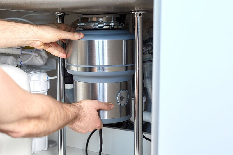 Hands installing a stainless steel garbage disposal under a kitchen sink, with water filters and pipes in the background.