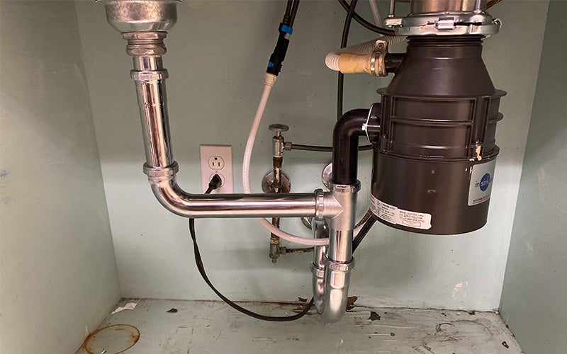Under-sink plumbing with a chrome drain pipe, P-trap, and a dark gray garbage disposal unit attached to a sink.