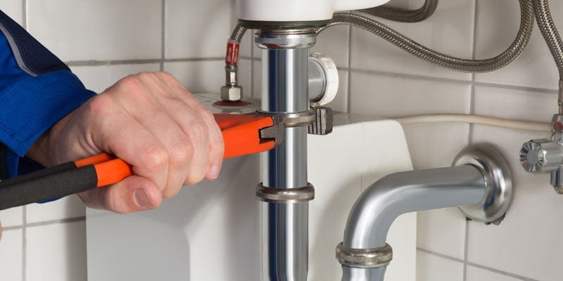 A plumber tightens a pipe fitting with an orange wrench while working on a sink.
