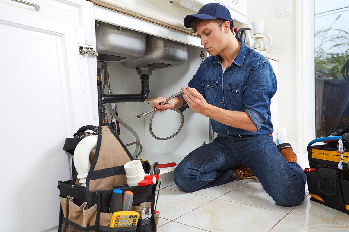 A plumber kneels and tightens a pipe under a sink with a wrench, surrounded by tools.