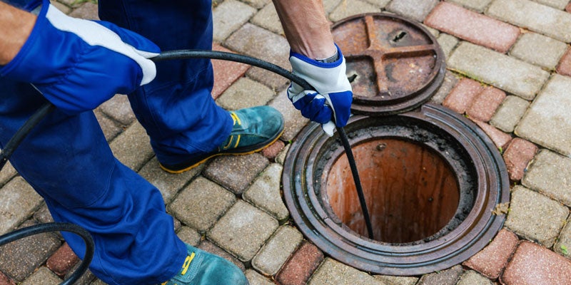 A worker in blue overalls inserts a flexible cable into a circular manhole cover set into a brick walkway.