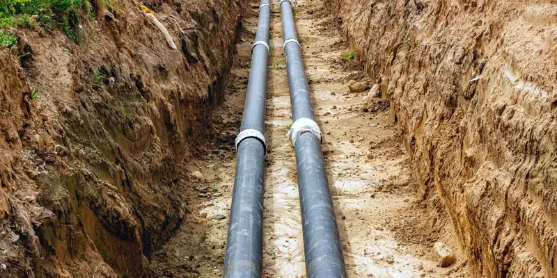 Two black pipes lie parallel within a freshly dug trench in dirt.