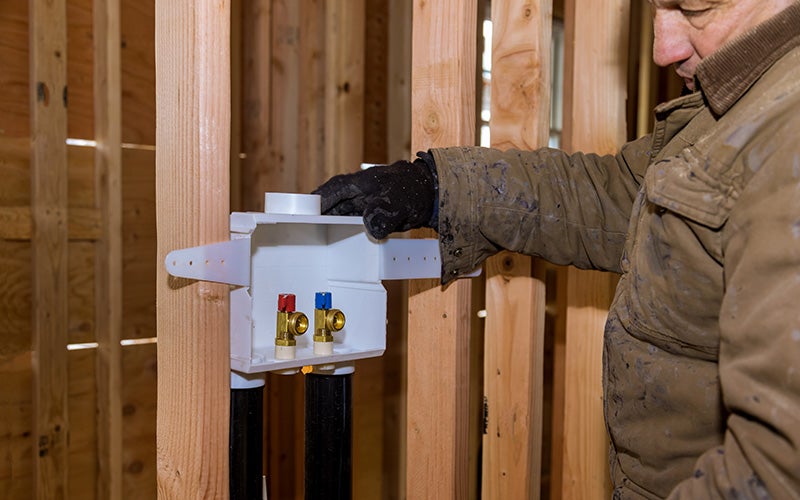 A worker installs a white washing machine outlet box with red and blue valves onto wooden wall studs.