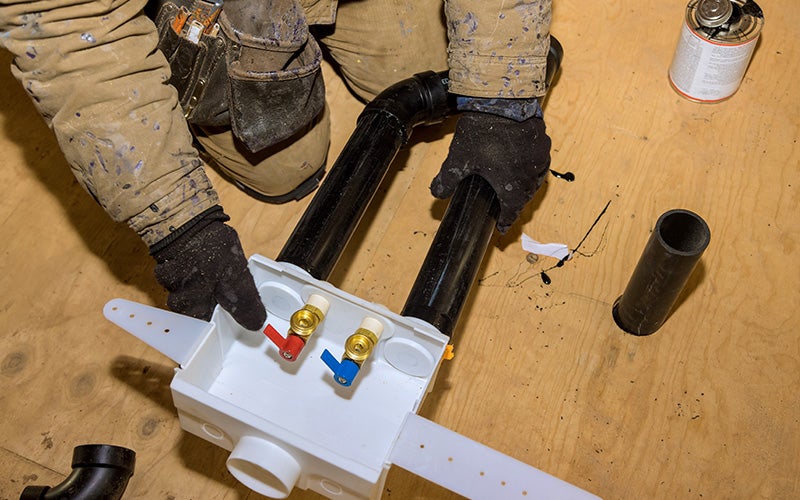 Plumber installing a white washing machine hookup box with red and blue valves and black PVC pipes on a wooden floor.