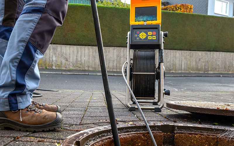 A person uses a pipe inspection camera with a screen to examine a drain cover on a sidewalk.