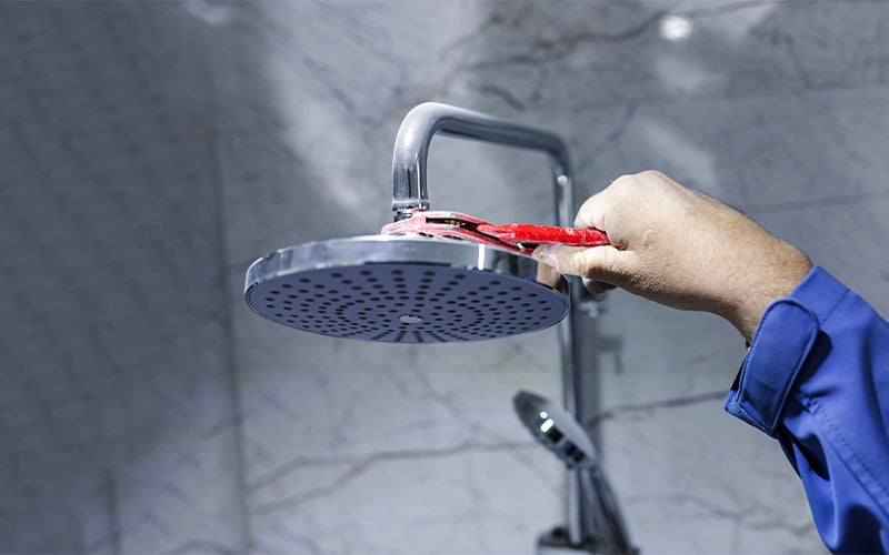 A person in a blue uniform adjusts a chrome showerhead against a tiled wall.