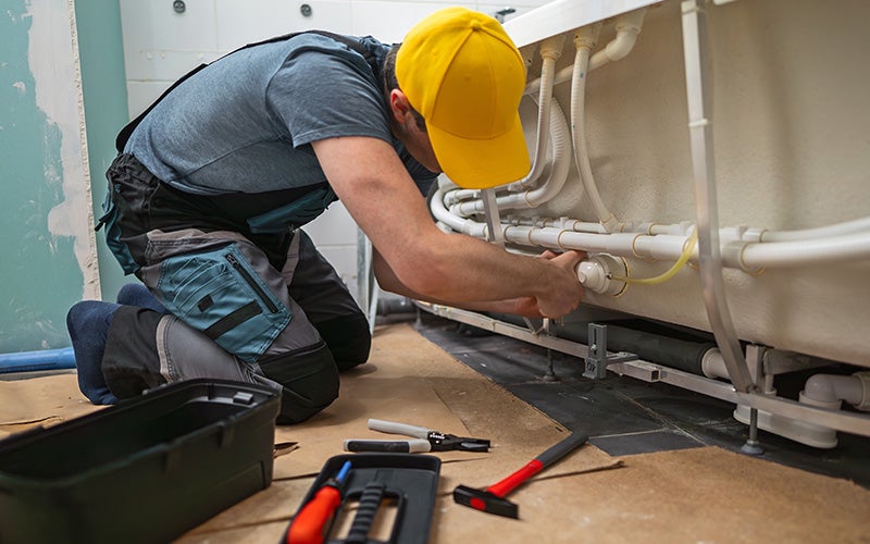 A plumber wearing a yellow cap works on pipes under a tub in a bathroom.