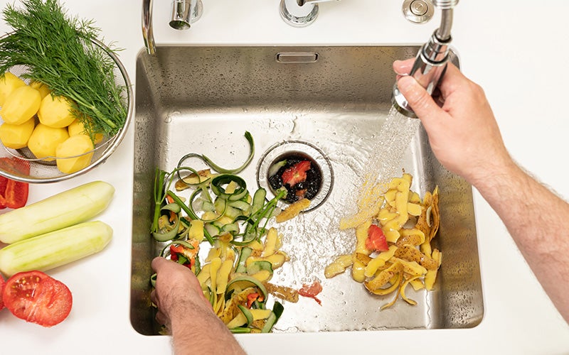 Vegetable scraps and peels are being discarded into a stainless steel kitchen sink.