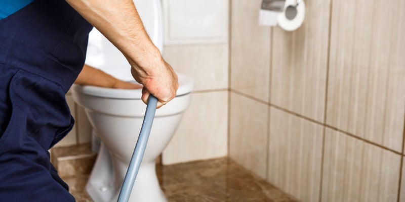 A plumber in blue overalls flushes a toilet in a tiled bathroom.