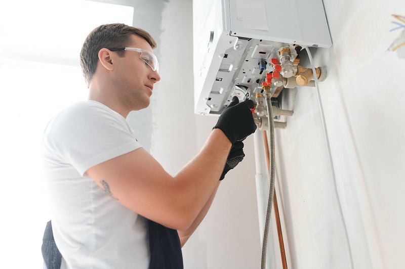 A man wearing safety glasses and gloves adjusts a wall-mounted boiler with copper pipes.