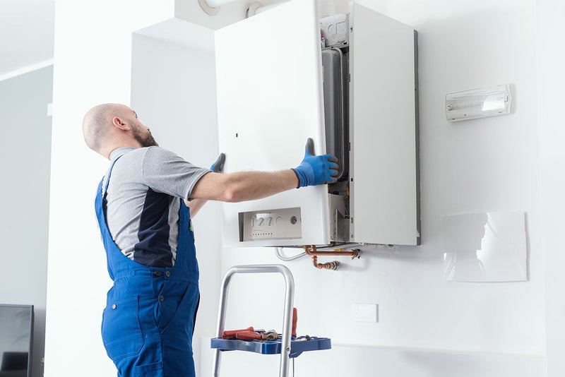 A plumber in blue overalls adjusts a white wall-mounted gas boiler.