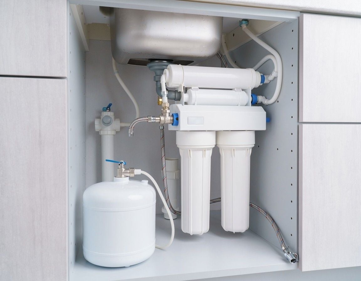 A water filtration system with pipes and cartridges sits inside a white kitchen cabinet.
