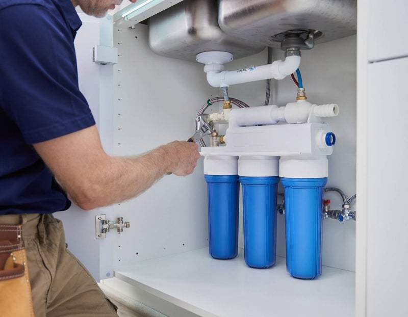 A plumber tightens a fitting on a water filtration system installed under a kitchen sink.